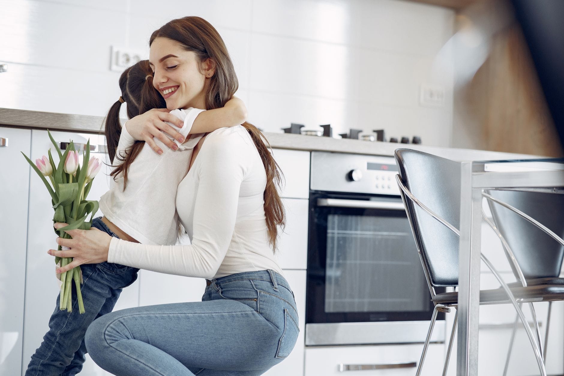 Mama przytula córkę w nowoczesnej kuchni, trzymając bukiet tulipanów Mama przytula córkę w nowoczesnej kuchni, trzymając bukiet tulipanów